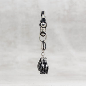 A silver-toned metal keychain or bag charm featuring a small carved pendant with black oxidized detailing, shown against a light gray textured background.