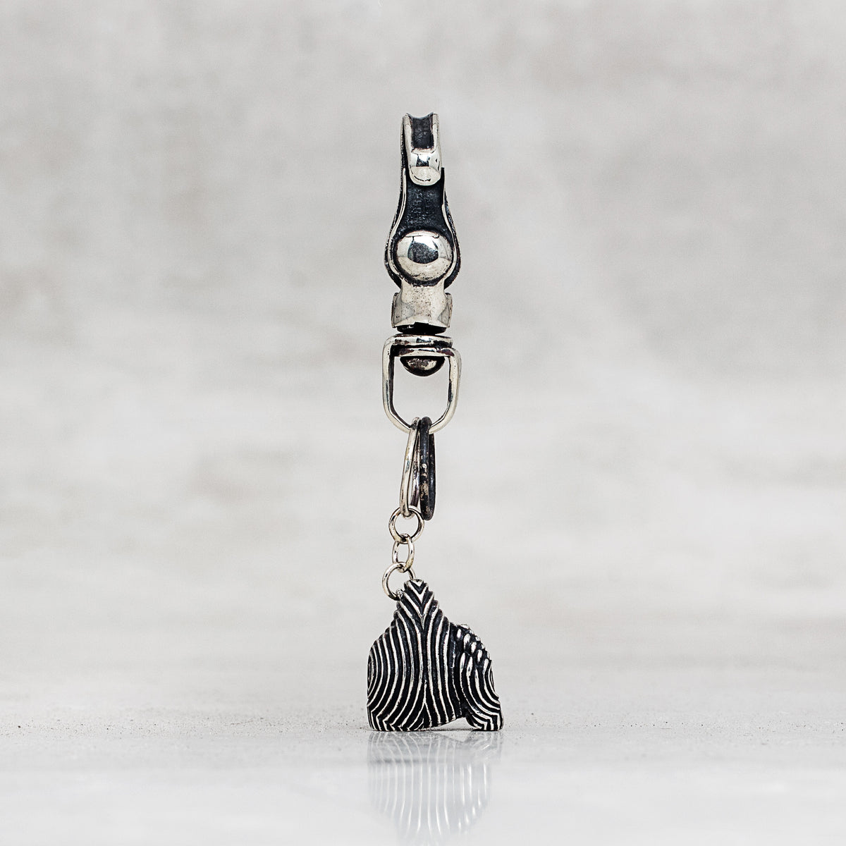 A silver-toned metal snap hook clasp with a dangling charm, shown against a light gray textured background.