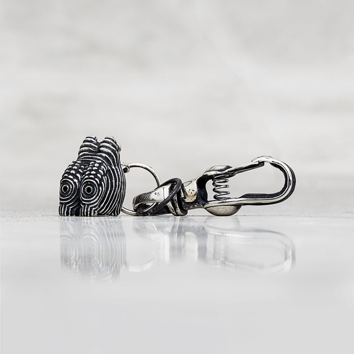 A sterling silver keychain featuring a stylized head charm with black and silver striped detailing, attached to a silver carabiner clip, displayed on a reflective light gray surface.