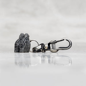 A sterling silver keychain featuring a stylized head charm with black and silver striped detailing, attached to a silver carabiner clip, displayed on a reflective light gray surface.