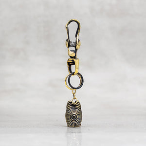 A gold and black metal keychain or bag charm with a lobster clasp, split ring, and a dangling pendant, shown on a light gray marble surface.
