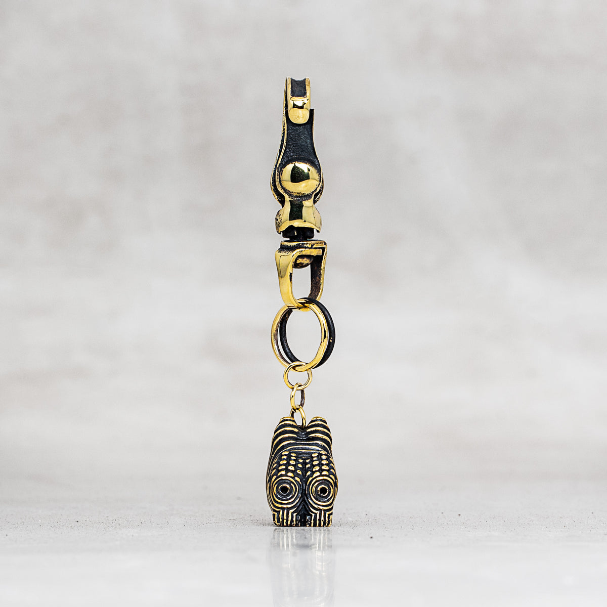 A gold and black metal accessory with a lobster clasp, jump ring, and a dangling charm, photographed against a light gray marble background.