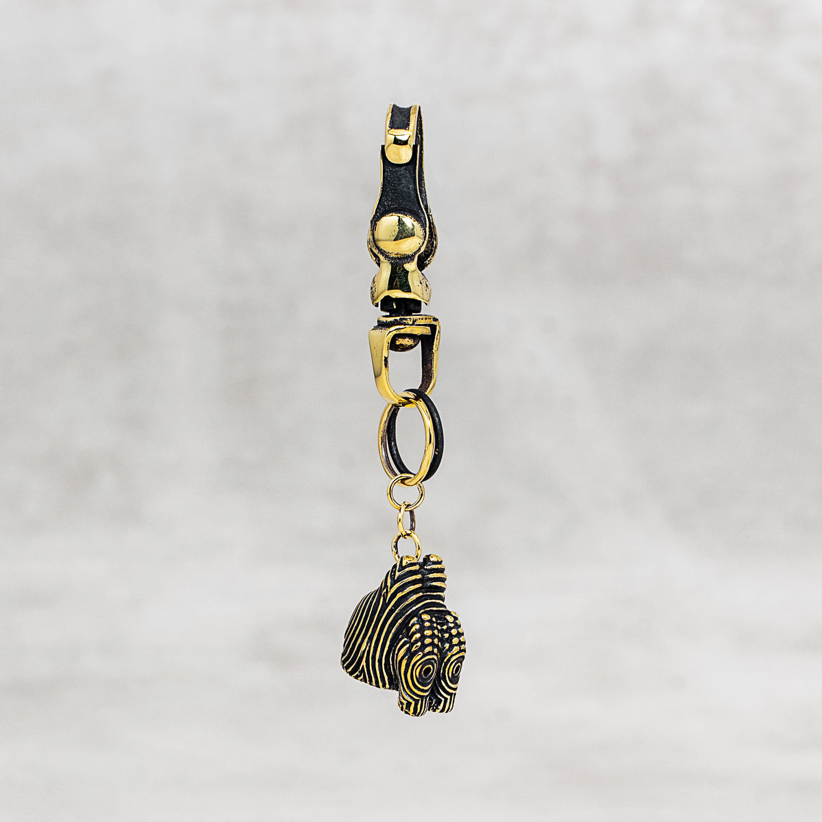 A gold-toned metal keychain or bag charm with a lobster clasp, ring connector, and a dangling textured ornament, shown against a soft light gray background.
