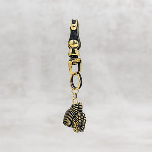 A gold-toned metal keychain or bag charm with a lobster clasp, ring connector, and a dangling textured ornament, shown against a soft light gray background.