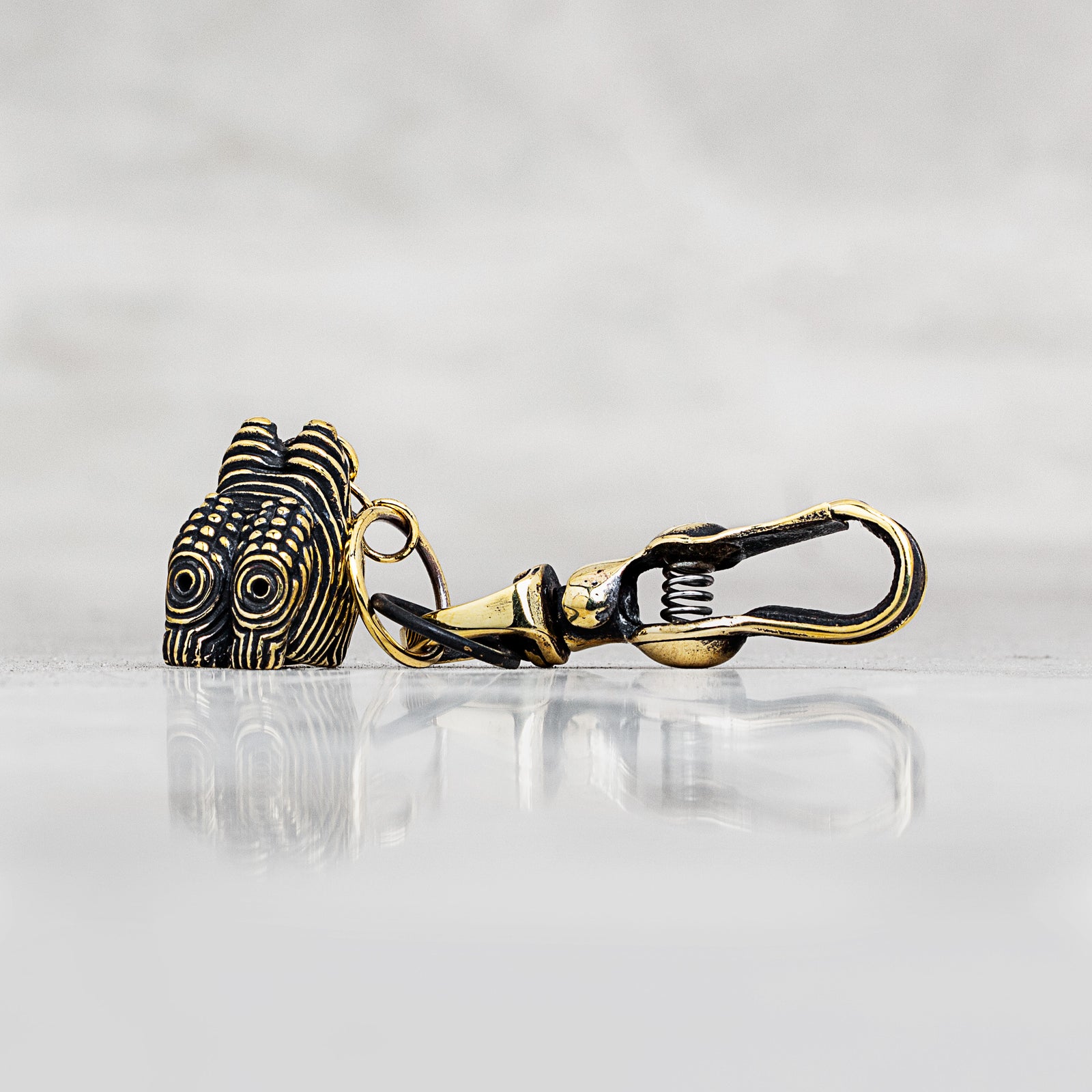 A close-up of a gold and black metal lobster clasp with a textured, patterned head, resting on a reflective light gray surface against a soft gray background.
