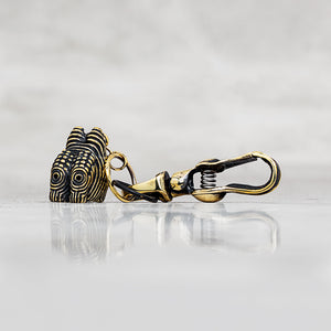 A close-up of a gold and black metal lobster clasp with a textured, patterned head, resting on a reflective light gray surface against a soft gray background.