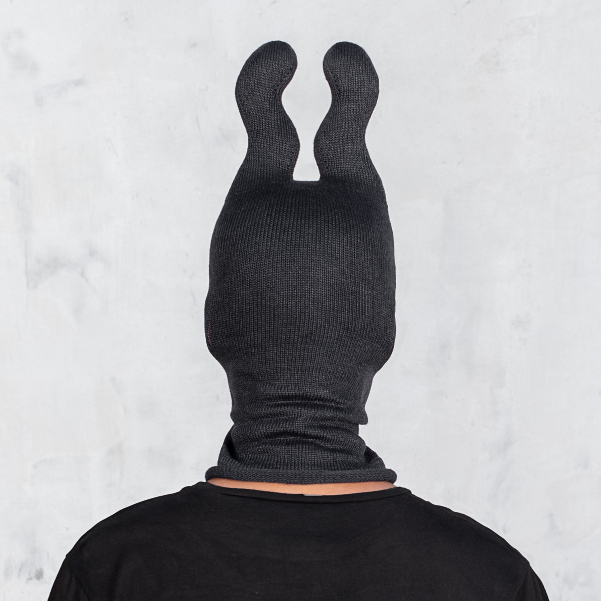 A person viewed from the back wearing a black ribbed balaclava-style head covering with two upright wavy ears, against a light gray textured wall.
