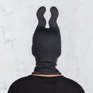 A person viewed from the back wearing a black ribbed balaclava-style head covering with two upright wavy ears, against a light gray textured wall.