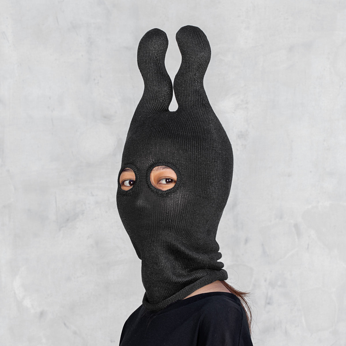 A person wearing a black ribbed knit  mask with two upright wavy ears and eye cutouts, shown against a light gray textured wall.