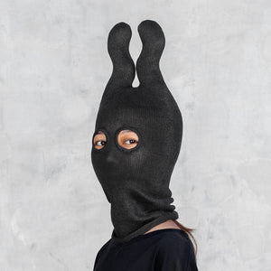 A person wearing a black ribbed knit  mask with two upright wavy ears and eye cutouts, shown against a light gray textured wall.