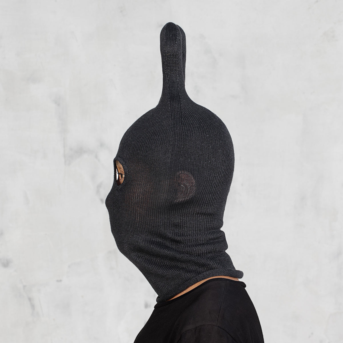 A person shown in side profile wearing a black ribbed knit hood with a tall pointed top and eye cutouts, against a light gray textured wall.