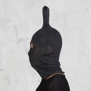A person shown in side profile wearing a black ribbed knit hood with a tall pointed top and eye cutouts, against a light gray textured wall.