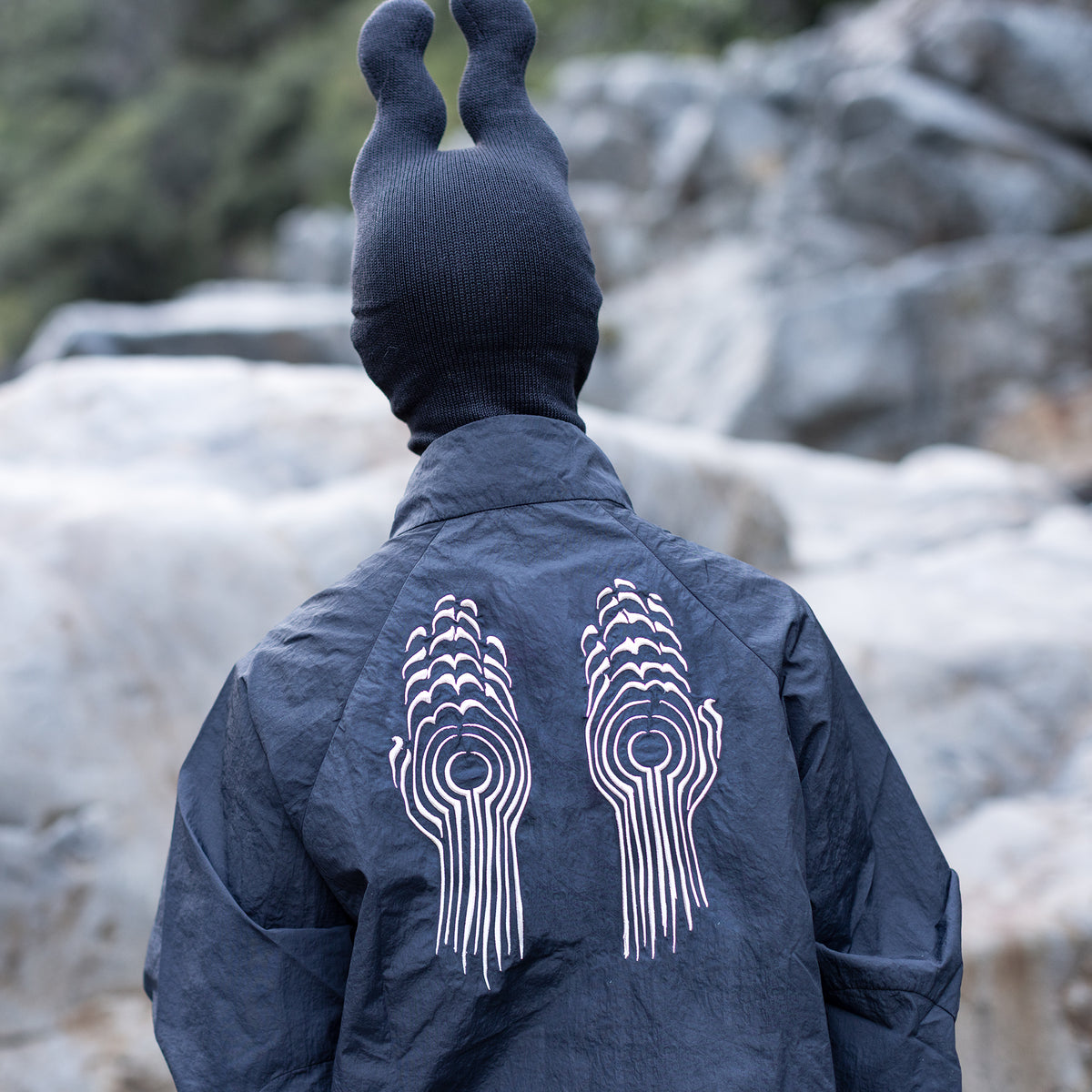 A person viewed from behind wearing a dark jacket with two large white line-art hand graphics on the back and a matching dark knit hood with rabbit-like ears, standing outdoors in front of pale rocks.