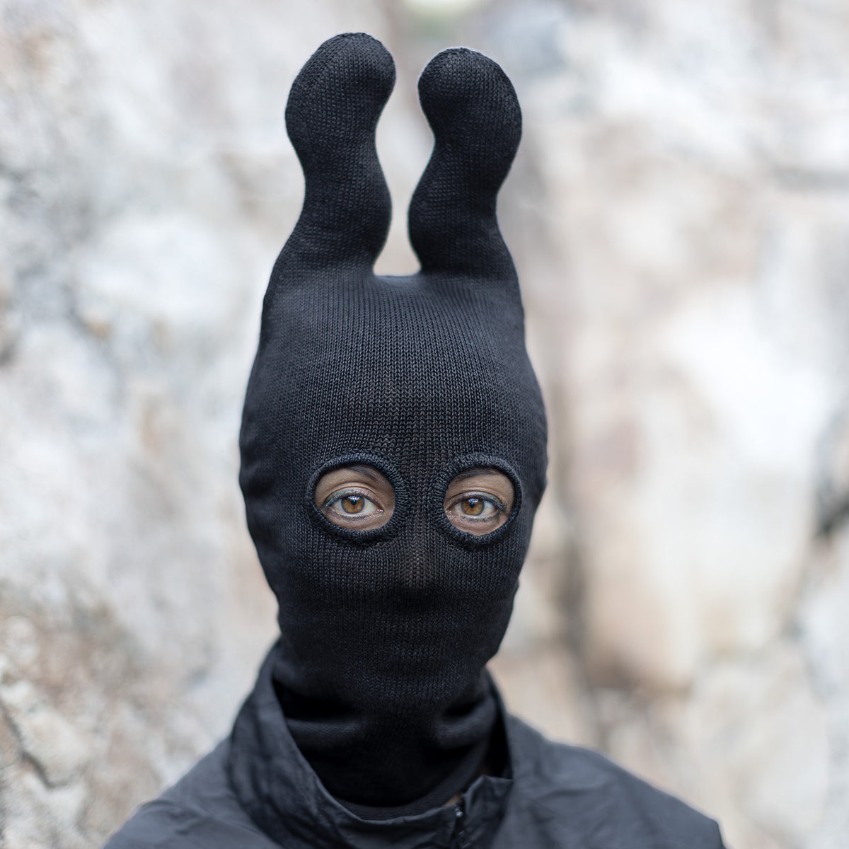 Person wearing a black knit balaclava with wavy upright horns in front of a beige blurred background