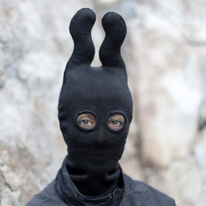 Person wearing a black knit balaclava with wavy upright horns in front of a beige blurred background