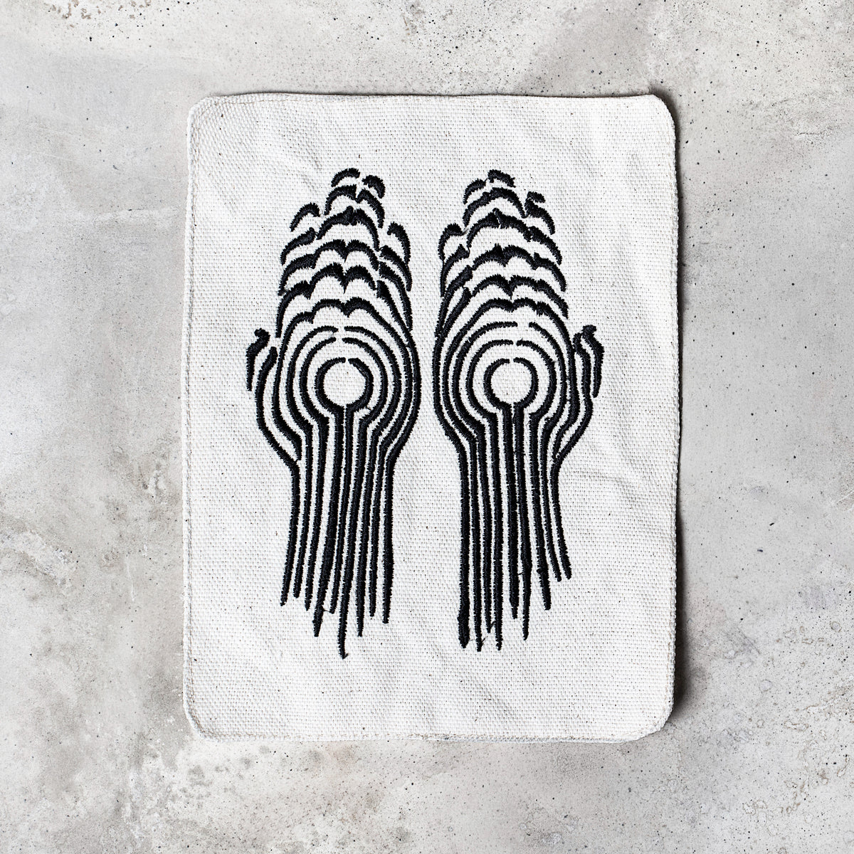 A white rectangular canvas patch with rounded corners, featuring a black graphic print of two stylized hands forming a circular motif, laid flat on a light gray textured surface.