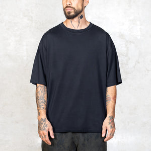 A tattooed person from the waist up facing front wearing a boxy cut black short sleeve tee. 