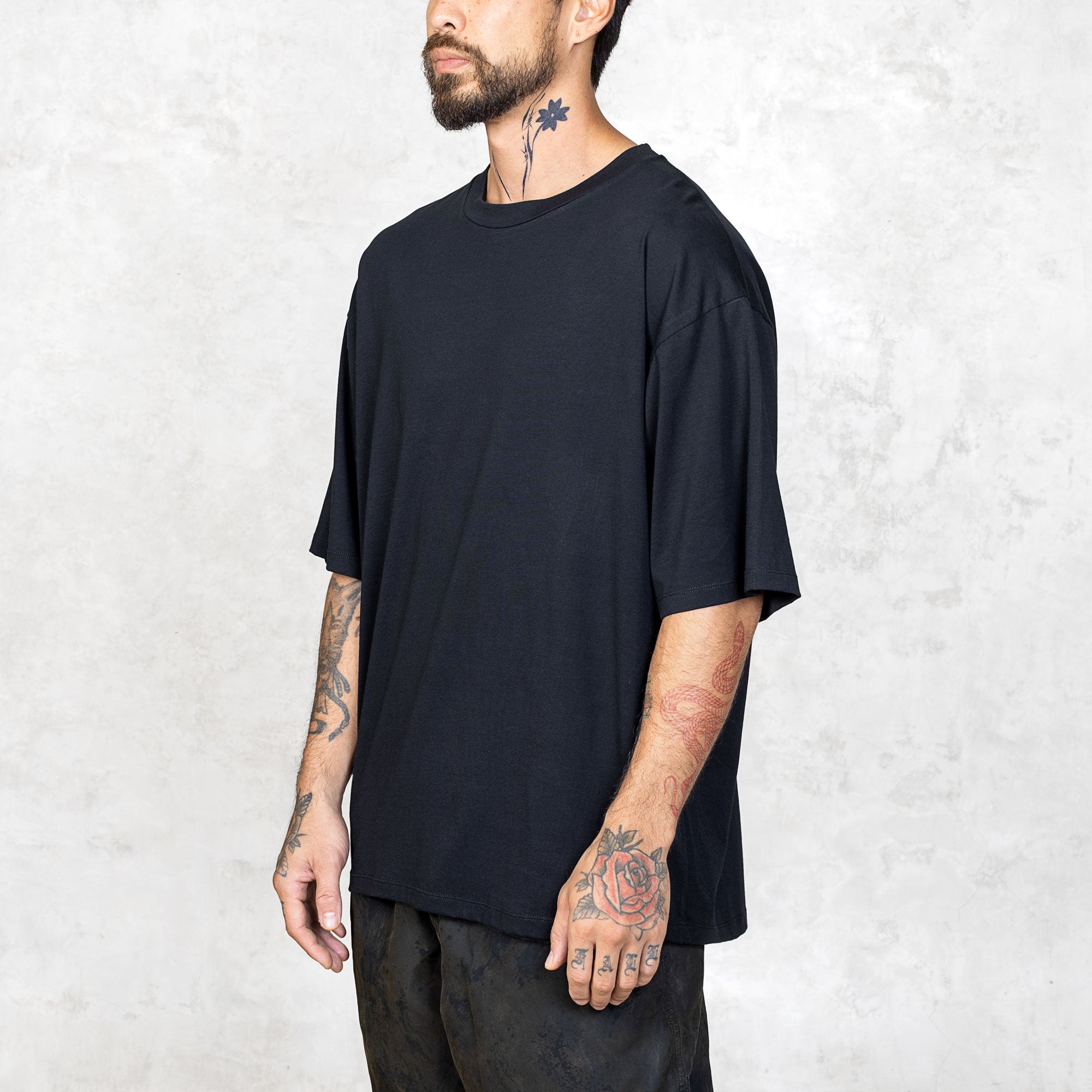 A tattooed person from the waist up wearing a boxy cut black short sleeve tee. 