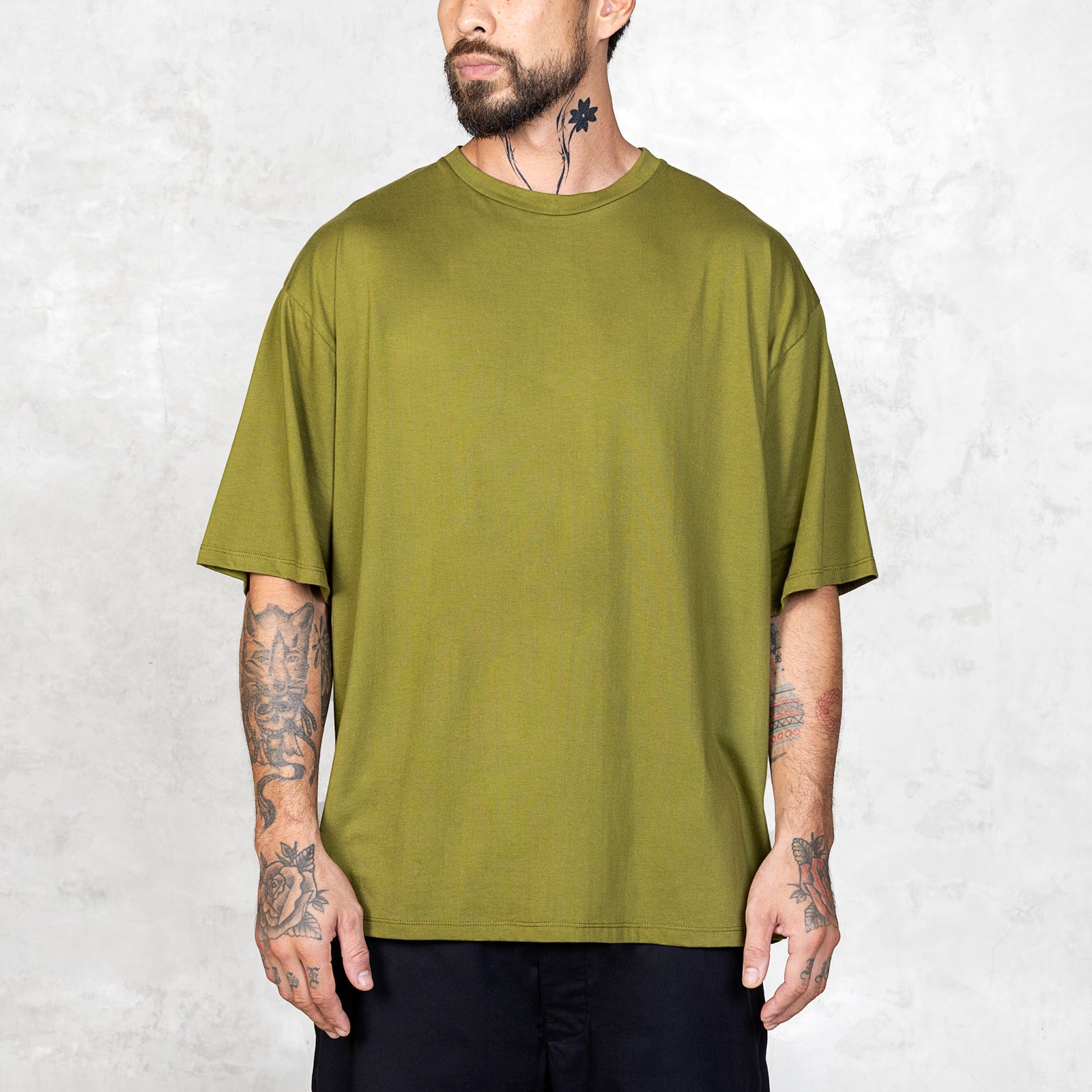 Tattooed person facing forward wearing an oversized green t-shirt against a light gray background