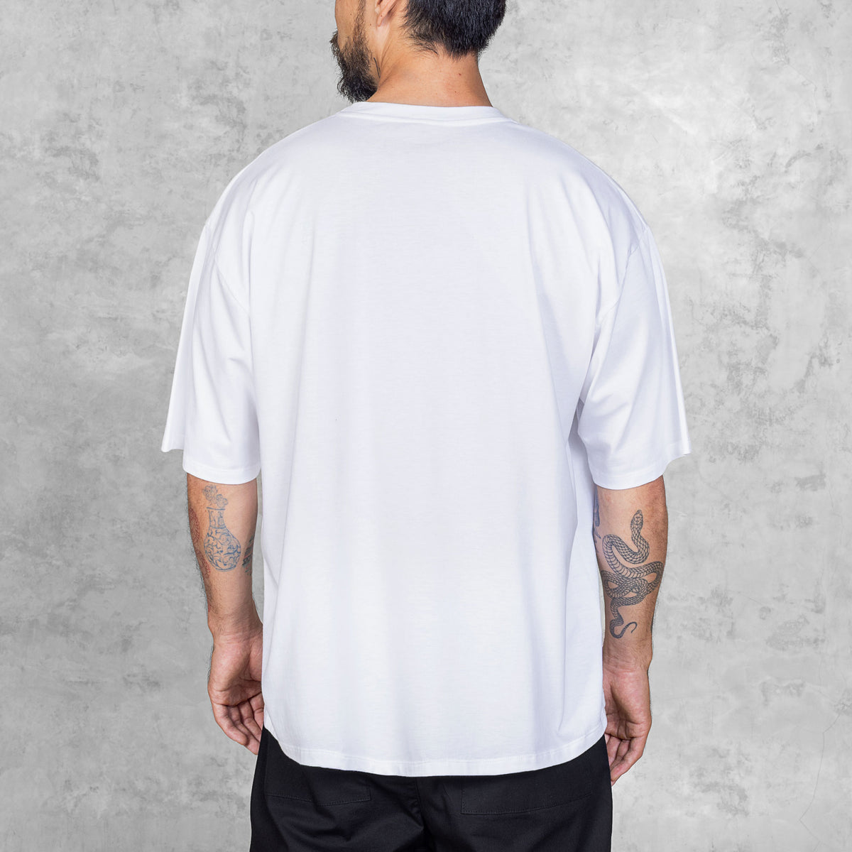 Tattooed person standing facing back from waist to head wearing black pants and a plain white boxy-style tee