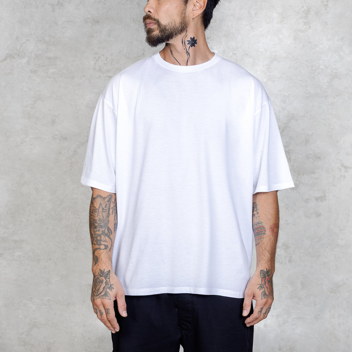 Tattooed person standing from waist to head facing front wearing black pants and a plain white boxy-style tee