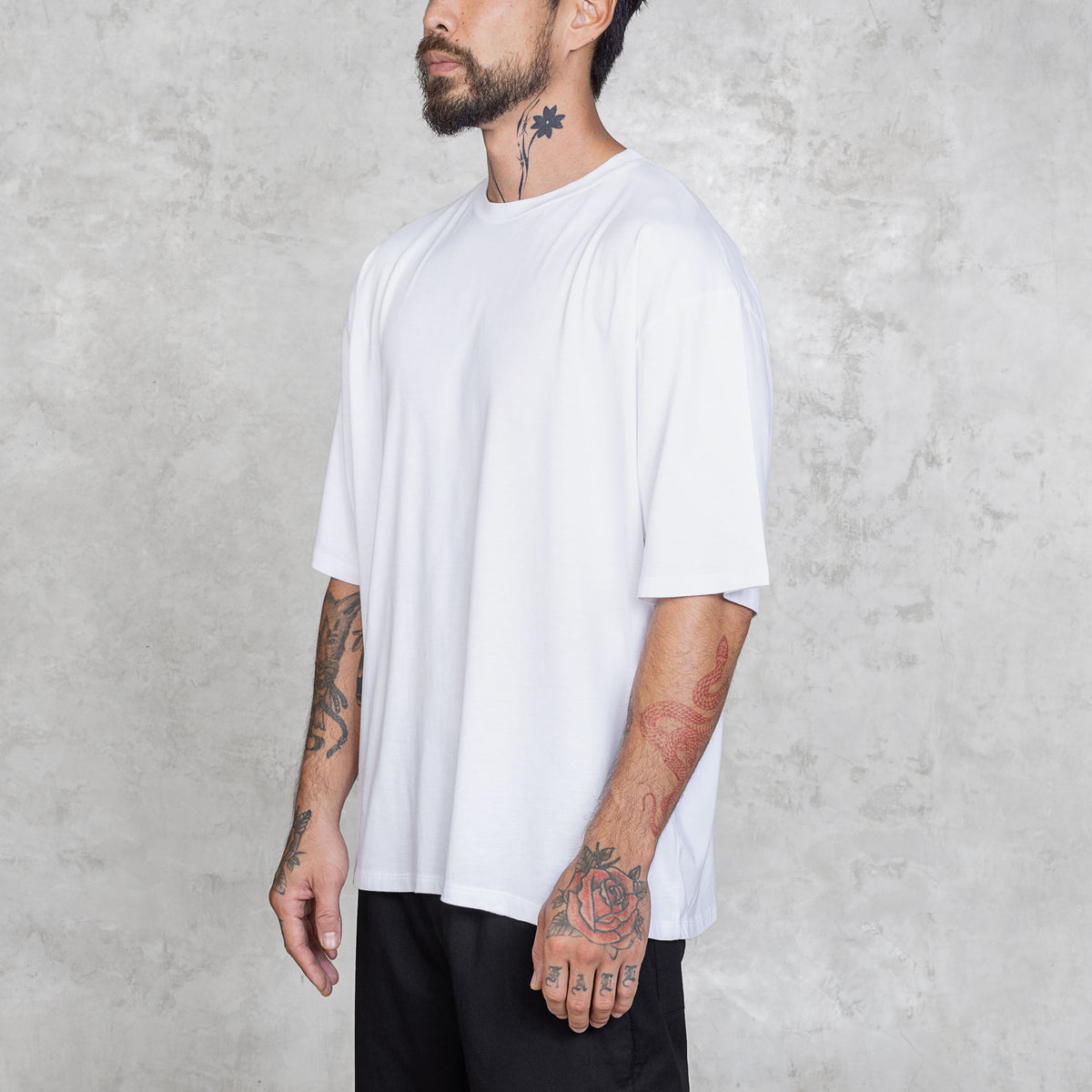 Tattooed person standing from waist to head wearing black pants and a plain white boxy-style tee