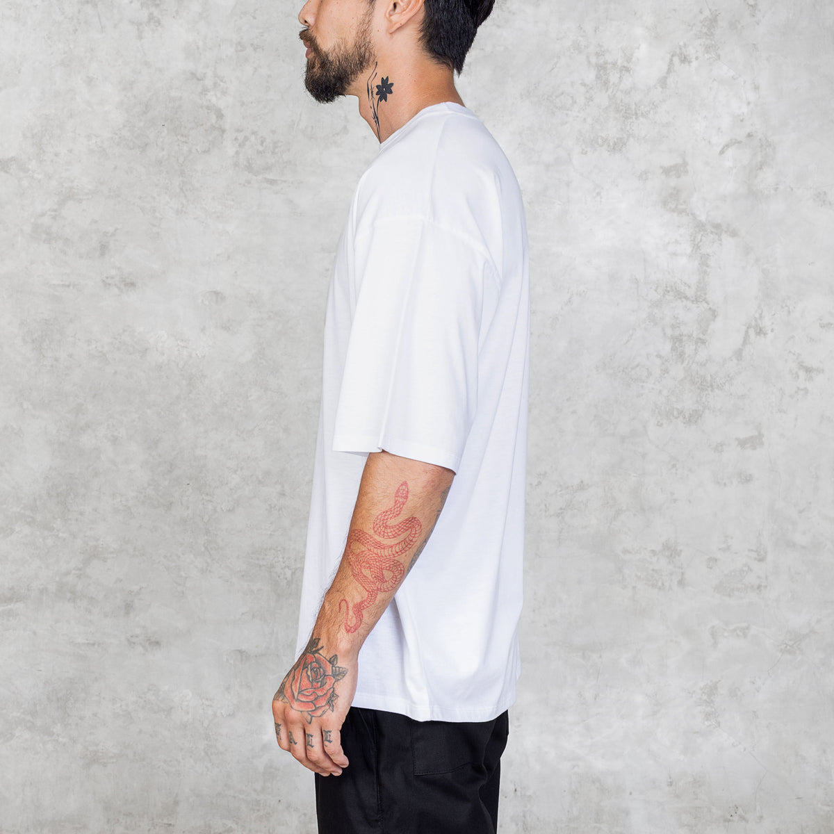 Tattooed person standing from waist to head wearing black pants and a plain white boxy-style tee