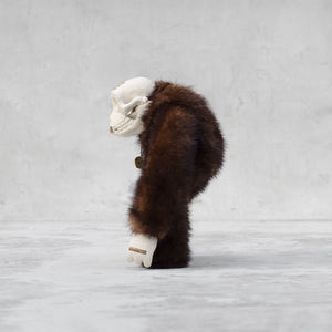 Side view of a ceramic, fur, and brass monkey skull figurine standing against a light gray background