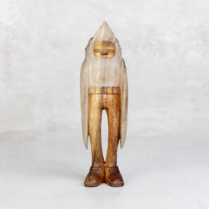 Wooden sculpture of a figure with a beak hood and wing arms on a white background