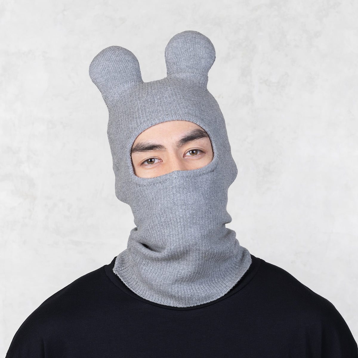 Person wearing a gray knit balaclava with animal ears on a light gray background