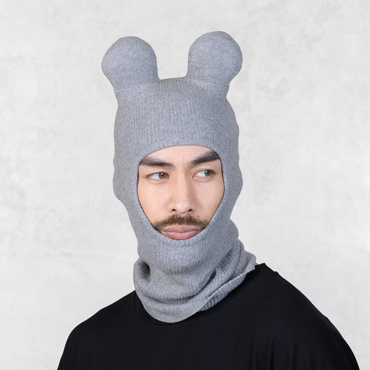 Person wearing a gray knit balaclava with bear ears on a light gray background