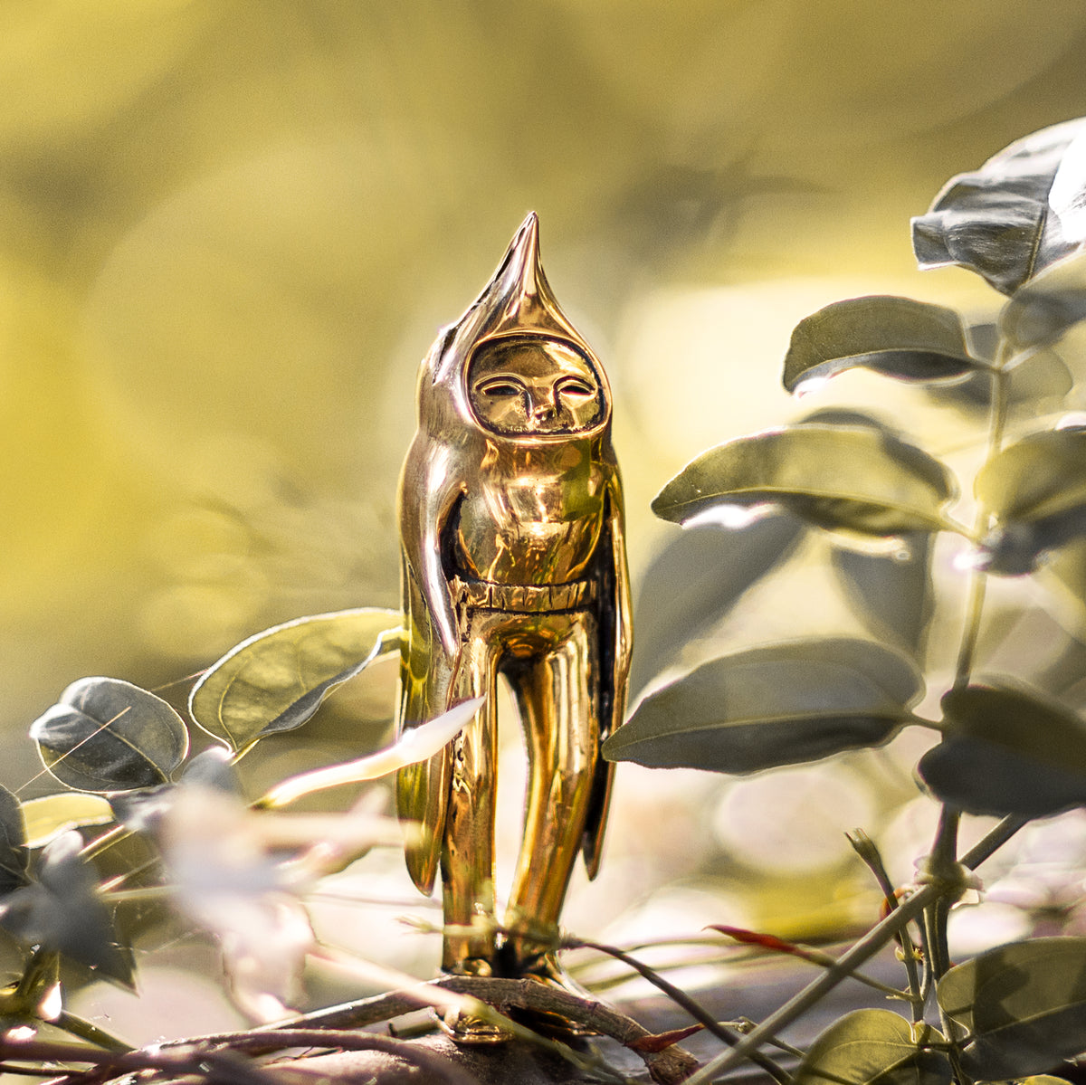 Golden brass statuette of a bird person standing amongst green leaves with a natural background