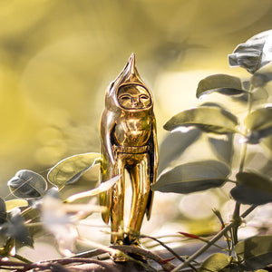 Golden brass statuette of a bird person standing amongst green leaves with a natural background