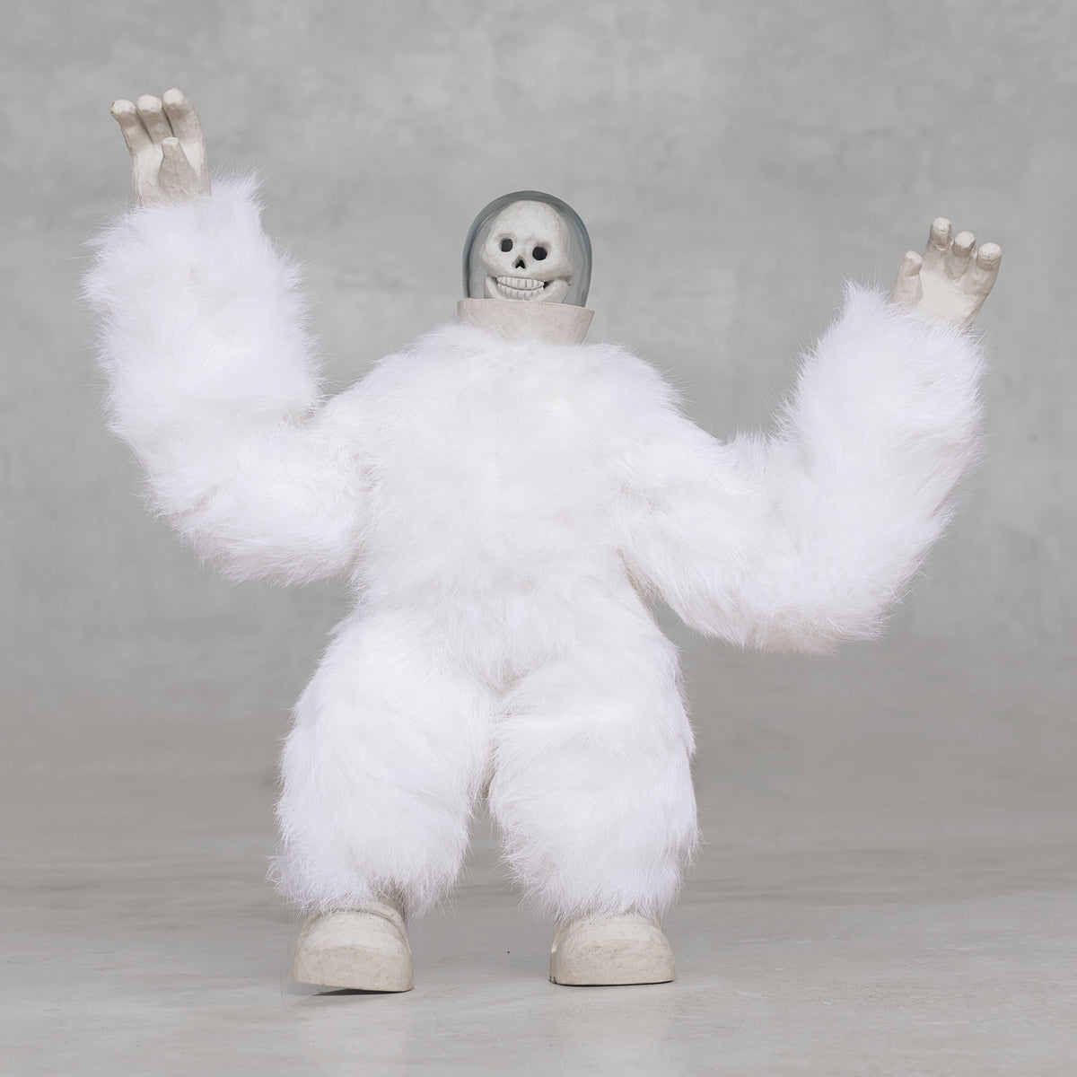 White furry creature statue with skull head with its arms raised against a light gray background 