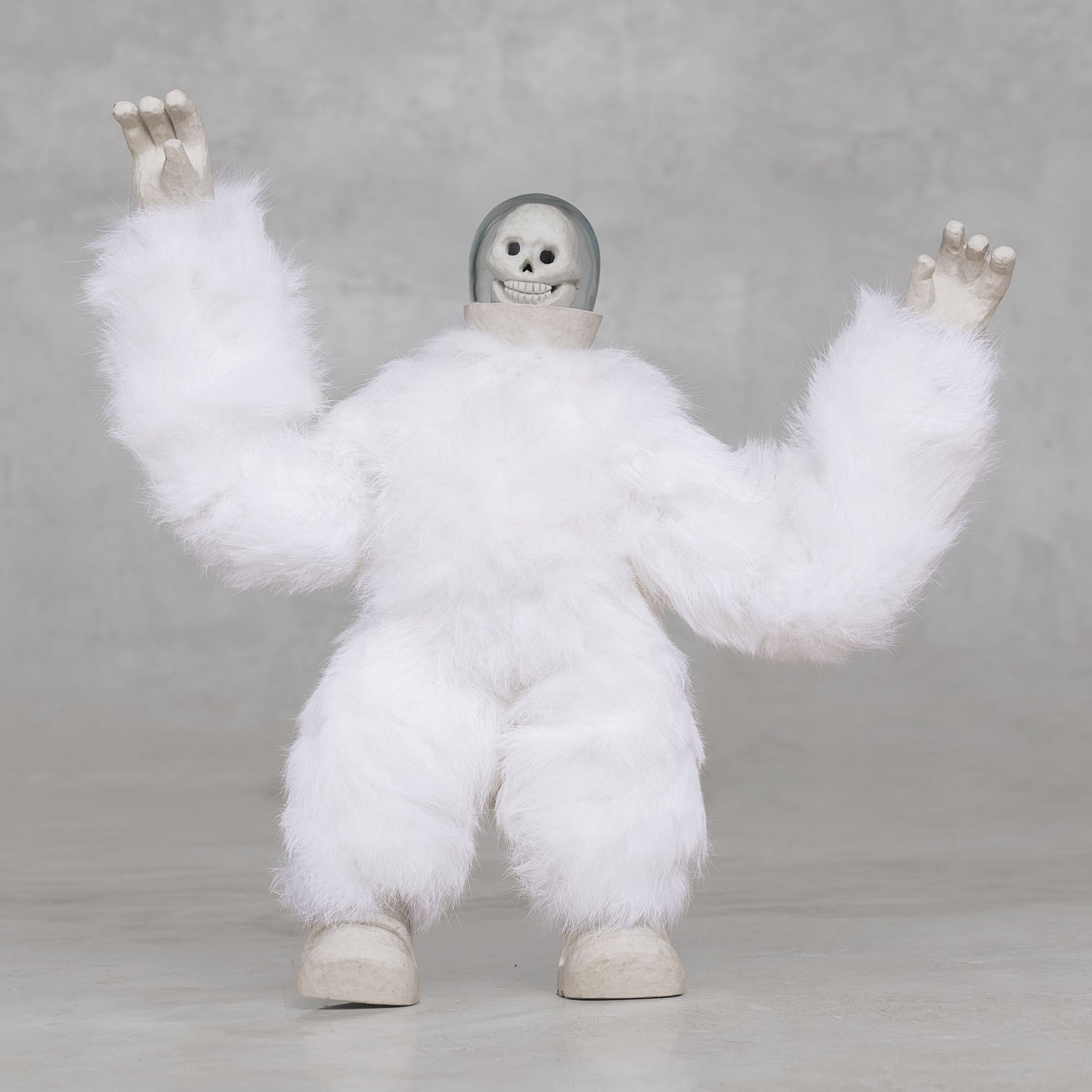 White furry creature statue with skull head with its arms raised against a light gray background 