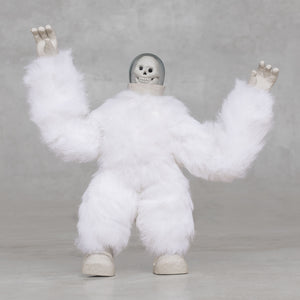 White furry creature statue with skull head with its arms raised against a light gray background 