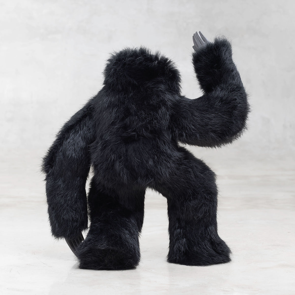 Back view of a black furry creature with one arm raised on a light gray background 