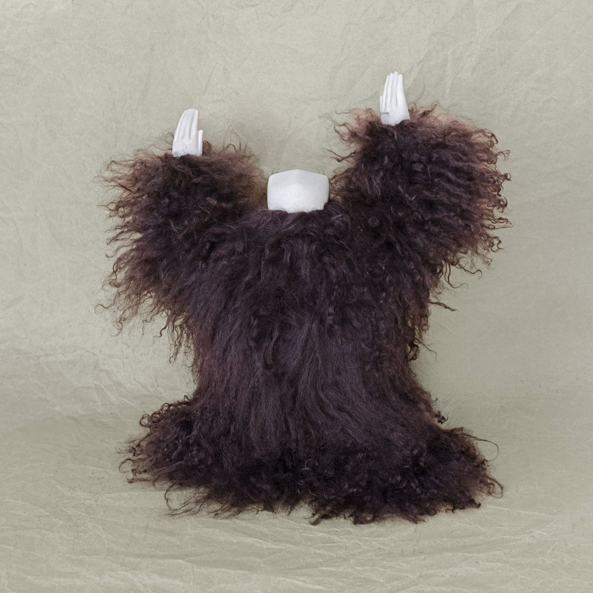 Back view of a creature sculpture made of ceramic and with its arms raised above its head against a beige background