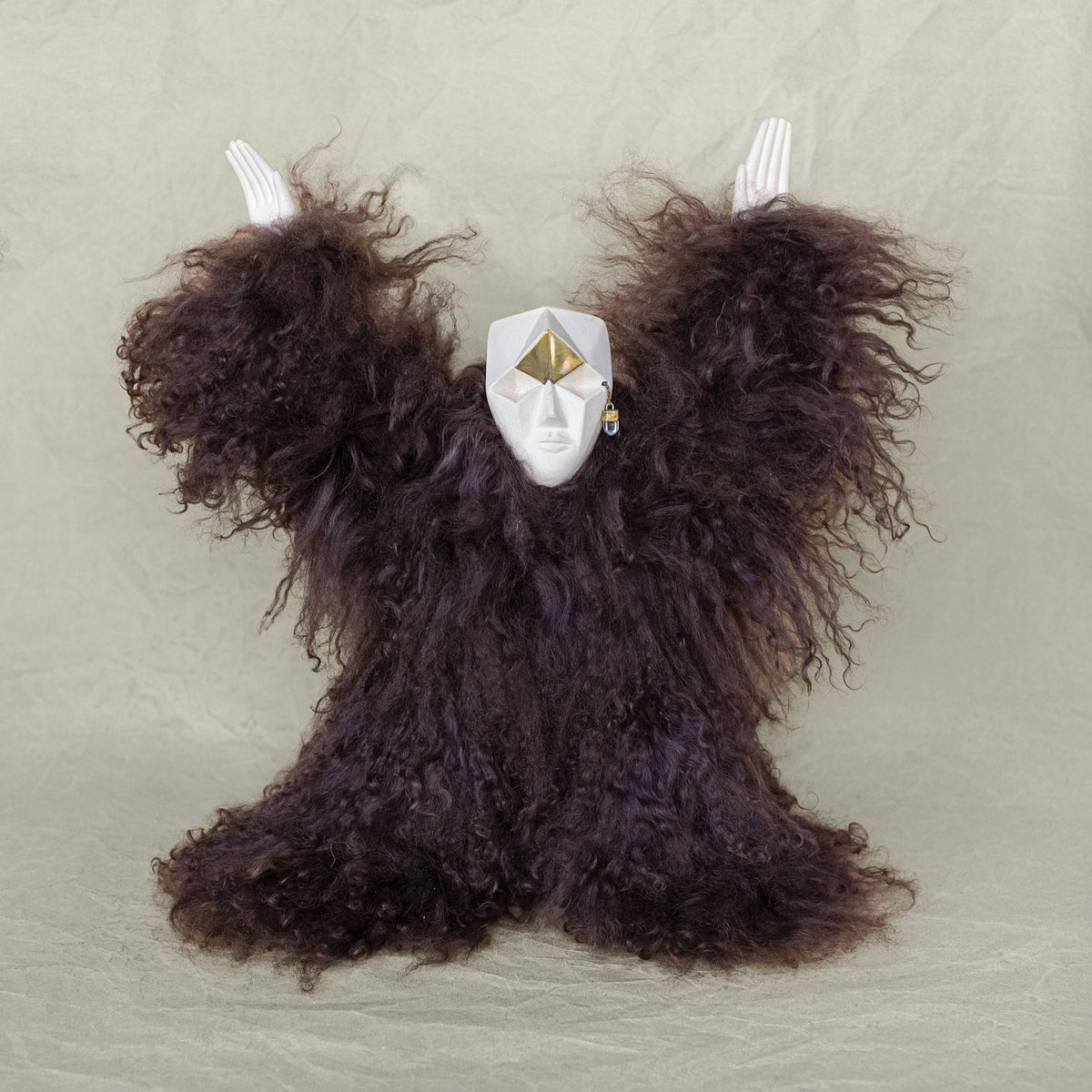 A creature sculpture made of ceramic, fur, and brass with its arms raised above its head against a beige background