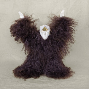 A creature sculpture made of ceramic, fur, and brass with its arms raised above its head against a beige background