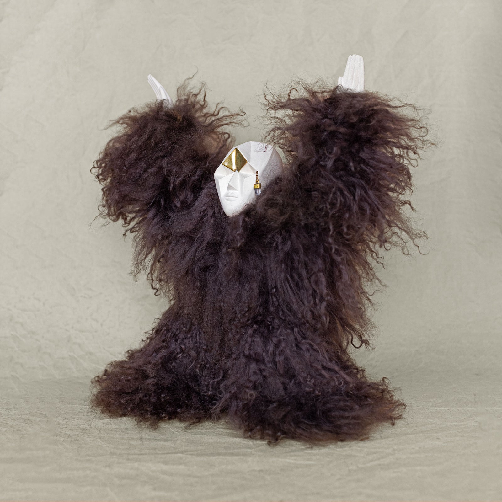 Quarter view of a creature sculpture made of ceramic, fur, and brass with its arms raised above its head against a beige background