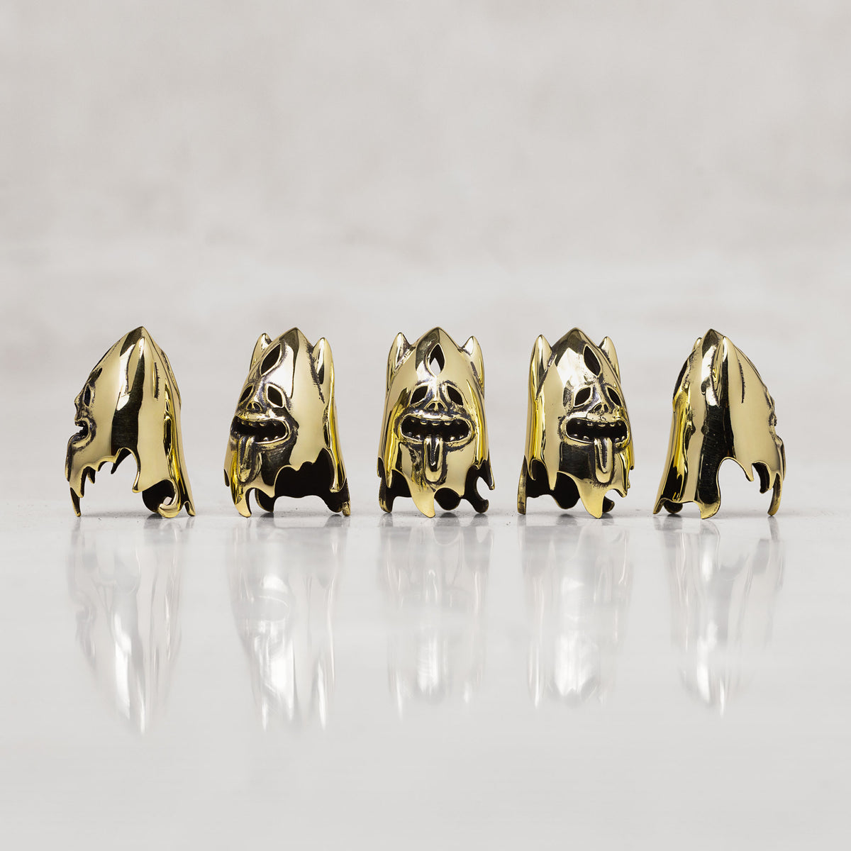 Five gold-toned skull-shaped thimbles stand arranged in a row on a reflective white surface against a light gray background.