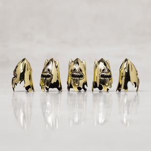 Five gold-toned skull-shaped thimbles stand arranged in a row on a reflective white surface against a light gray background.