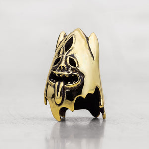 A gold-toned metal thimble shaped like a stylized ghostly face with a protruding tongue, photographed on a light gray reflective surface.