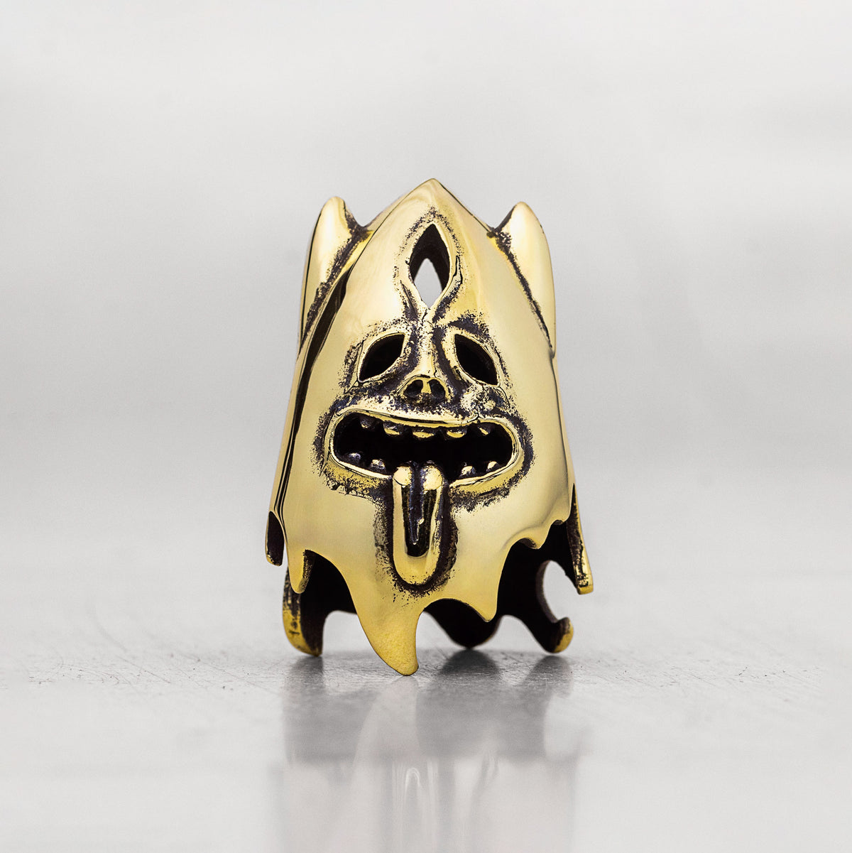 A gold-toned metal thimble shaped like a stylized ghost face with a protruding tongue and dark recessed details, photographed on a light gray reflective surface.