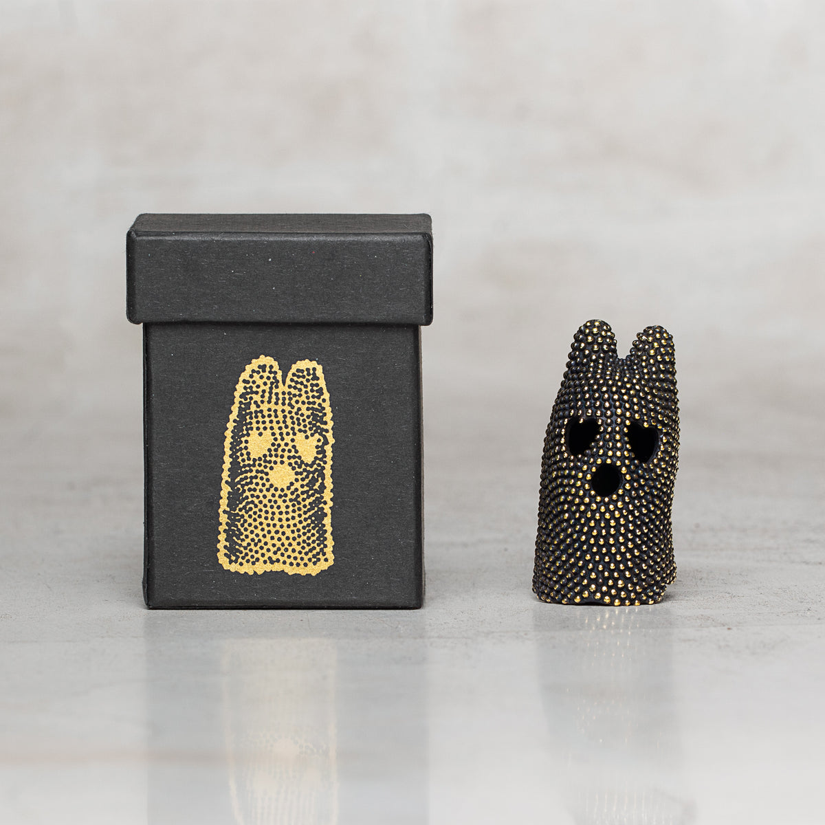 A small black speckled figurine with heart-shaped eyes and a black box featuring a gold dotted illustration, set against a light marble background.