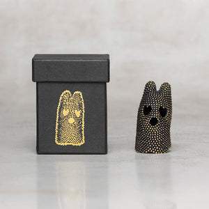 A small black speckled figurine with heart-shaped eyes and a black box featuring a gold dotted illustration, set against a light marble background.