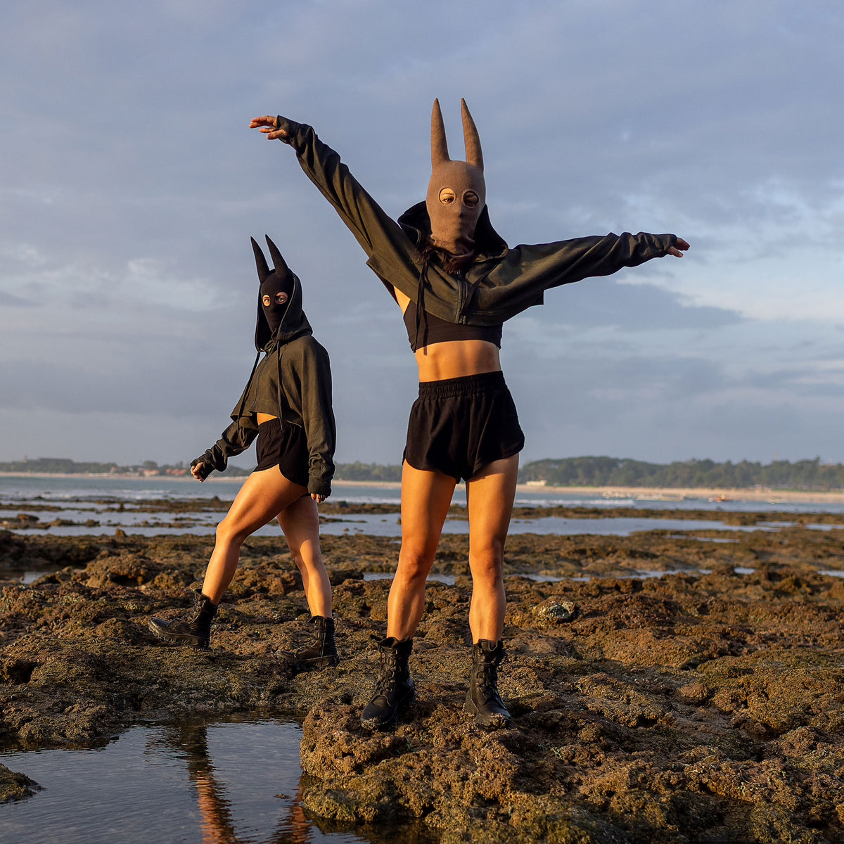 Two people wearing spire masks, cropped green hoodies, black shorts, and black boots on rocks by the ocean. The person on the left is walking to the left looking at the camera and the person on the right is looking at the camera with their arms raised. 