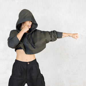 Person facing forward from head to knees punching to the right side wearing a green and black patterned cropped hoodie with the hood up to cover their face against a light gray background