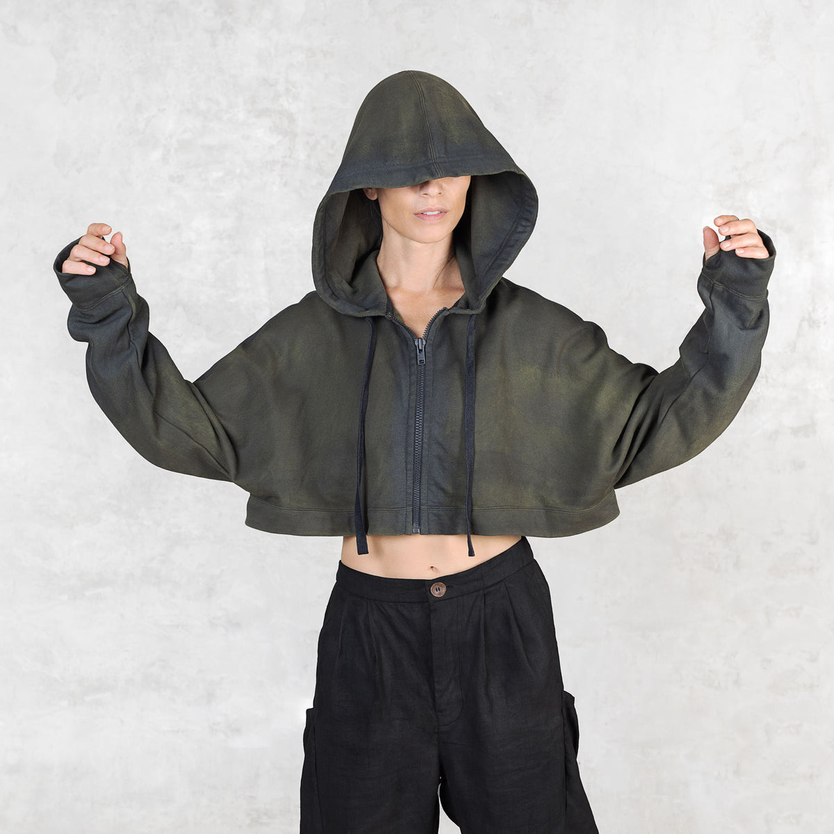 Person facing forward from head to knees with their arms slightly raised wearing a green and black patterned cropped hoodie with the hood up to cover their face against a light gray background